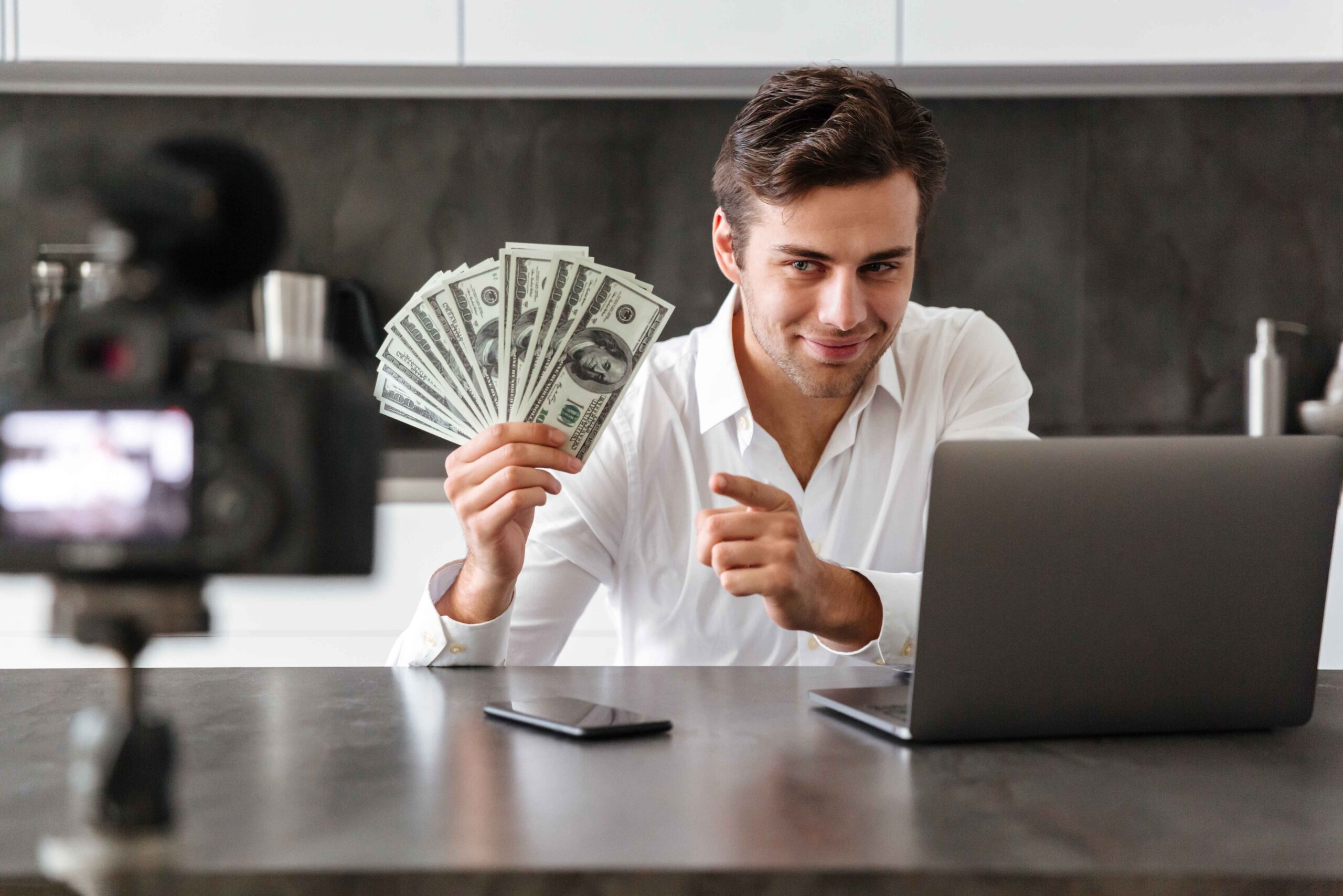 Young man recording a video blog at home while showing US dollar bills and using a laptop